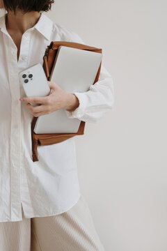 Young pretty woman in white linen shirt holding mobile phone, laptop computer and office documents over white wall