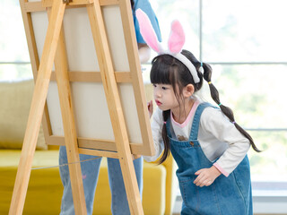 Portrait shot of smiling young asian woman pretty mother helping little cute daughter girl paint picture on canvas having fun together and spending family leisure time in living room at home.