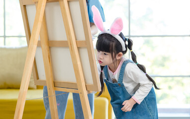 Portrait shot of smiling young asian woman pretty mother helping little cute daughter girl paint picture on canvas having fun together and spending family leisure time in living room at home.