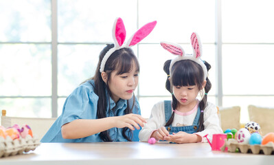 Asian cute little children girl wearing funny bunny ears headbands and young happy mother smile decorating painting eggs while sitting together in living room table family preparing for Easter holiday