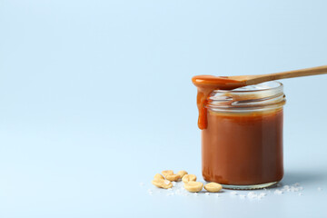 Jar of tasty salted caramel, spoon and peanuts on light blue background, space for text