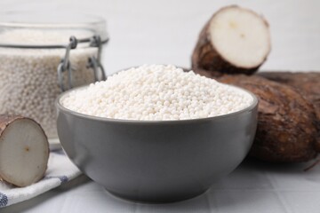 Tapioca pearls in bowl and cassava roots on white tiled table, closeup