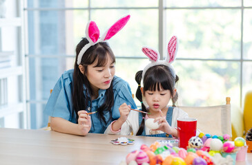 Asian cute little children girl wearing funny bunny ears headbands and young happy mother smile decorating painting eggs while sitting together in living room table family preparing for Easter holiday