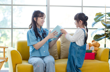 Happy mother's day Asian little cute girl child congratulating mom. Young mother and daughter smiling and holding gift on yellow cozy sofa in living room at home, Family holiday and togetherness.