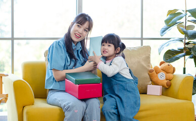 Happy mother's day Asian little cute girl child congratulating mom. Young mother and daughter smiling and holding gift on yellow cozy sofa in living room at home, Family holiday and togetherness.