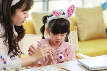 Asian young happy mother and little cute children daughter girl wearing funny bunny ears headbands with painting eggs while sitting together on table have laptop computer. Family celebrating Easter.