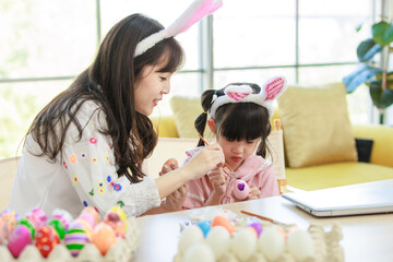 Obraz na płótnie Canvas Asian young happy mother and little cute children daughter girl wearing funny bunny ears headbands with painting eggs while sitting together on table have laptop computer. Family celebrating Easter.