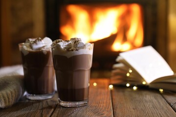Glasses with hot cocoa, marshmallows, lights and book on wooden table near fireplace