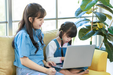 Asian young mother and her little cute daughter girl learning study from laptop computer, sitting on yellow sofa in living room at home, Happy family, Education and early development concept.