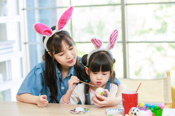 Asian cute little children girl wearing funny bunny ears headbands and young happy mother smile decorating painting eggs while sitting together in living room table family preparing for Easter holiday