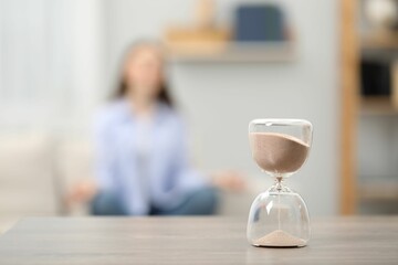 Hourglass with flowing sand on desk. Woman meditating indoors, selective focus