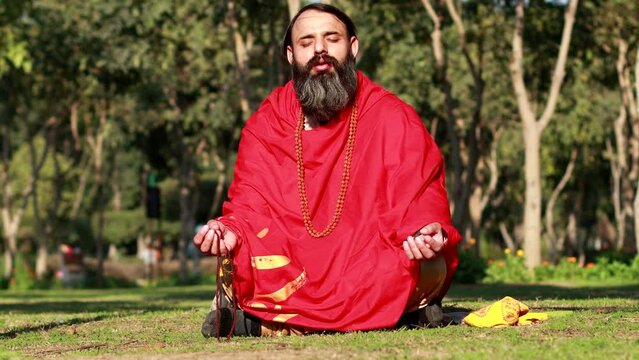 Beard yogi of Indian ethnicity chanting mantras sitting in lotus position outdoor in nature holding rudraksha in hands with eyes closed