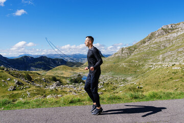 An athletic man jumps on a rope against the background of mountains. Sunny day, training outside.