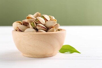 Tasty pistachios in bowl on white wooden table against olive background, closeup. Space for text