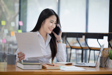 Happy asian young businesswoman using digital smartphone sitting in office working space, Asian female employee using laptop talking on the phone at workplace.
