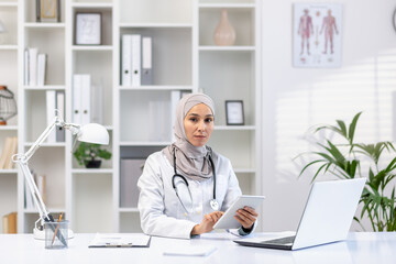 Professional Muslim female doctor in a hijab and white coat working at her office, using a tablet, with laptop and stethoscope on desk.