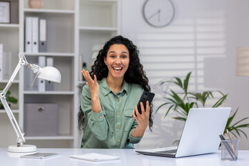 Enthusiastic mature Hispanic lady multitasking in a bright home office, working on laptop and...