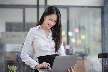 Happy asian young businesswoman using laptop with documents in office working space.