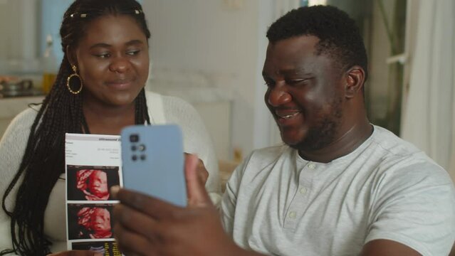Handheld shot of young black spouses calling parents on video on smartphone to show ultrasound image of unborn baby