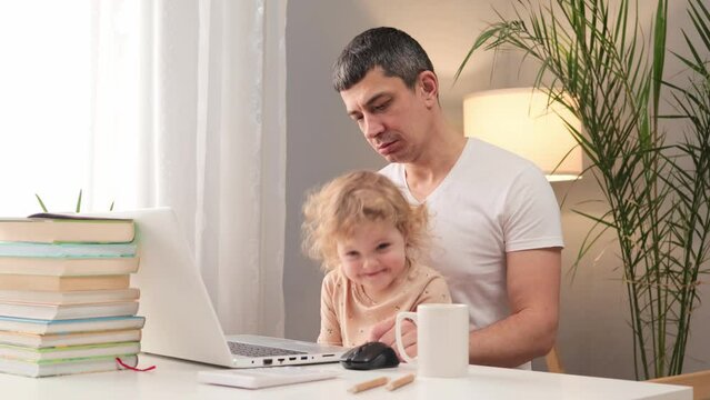 Disappointed sad unhappy man wearing white T-shirt working on laptop with his infant baby sitting at table at home having lots of work and childcare responsibilities