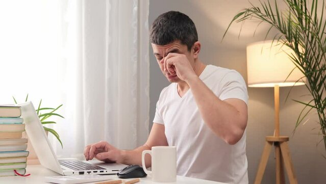 Tired exhausted Caucasian man wearing white t-shirt sitting working on laptop computer at home feeling sleepy and overworked rubbing his eyes feels fatigue from long hours online work