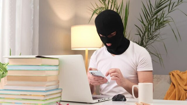 Unknown unrecognizable man in balaclava using somebodies laptop computer feeling free and relaxed on his dangerous work drinking coffee while finding stealing private information