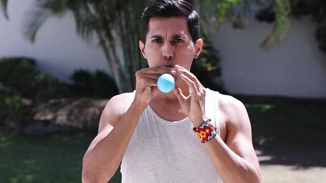 Serious young beardless man inflating a latex balloon in tropics