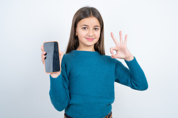 Young beautiful teen girl wearing blue T-shirt holding in hands cell showing ok-sign