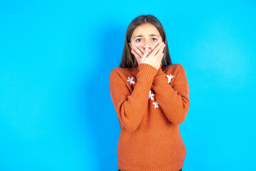 Stunned Young beautiful teen girl wearing orange knitted sweater covers both hands on mouth,...