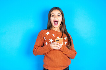 Young kid girl wearing orange sweater holds mobile phone in hands and rejoices positive news, uses...
