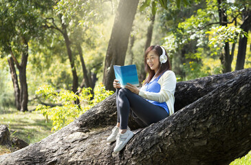 a young pleased asian woman wears headphones for listening music,sitting on a large spreading tree tree and reading a book