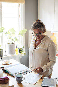 Mature woman photographing financial bills while standing in kitchen at home