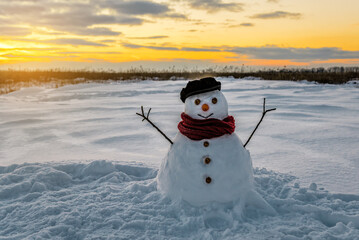 Cute snowman in cap and red scarf at sunset