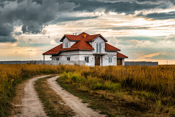 Abandoned farmhouse in the field at sunset under stormy clouds. Rural road