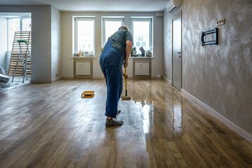 Professional worker lacquering varnishing parquet floors using roller. Home renovation