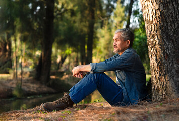 asian senior man with grey hair and beard sitting under a big tree in the forest park in the morning sun,mature male travel in nature,concept of elderly pensioner lifestyle,travel,camping,relaxation