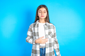Fototapeta na wymiar Beautiful kid girl wearing plaid shirt touches tummy, smiles gently, eating and satisfaction concept.