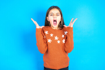 Crazy outraged beautiful kid girl wearing orange sweater screams loudly and gestures angrily yells...