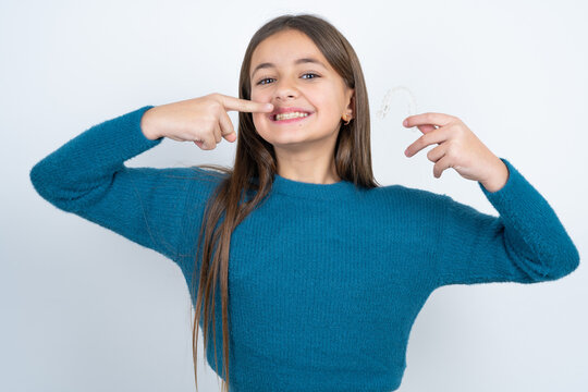 beautiful kid girl wearing blue sweater holding an invisible aligner and pointing perfect straight teeth. Dental healthcare and confidence concept.