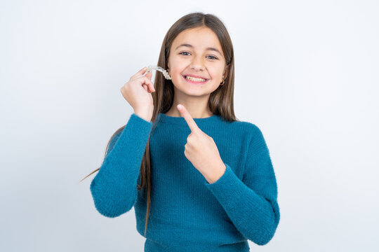 beautiful kid girl wearing blue sweater holding an invisible aligner and pointing at it. Dental healthcare and confidence concept.