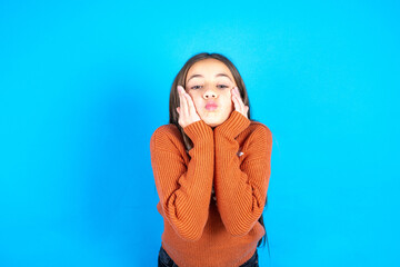 Young kid girl wearing orange knitted sweater with surprised expression keeps hands under chin keeps lips folded makes funny grimace