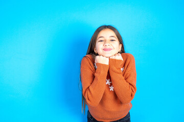 Young kid girl wearing orange knitted sweater holds hands under chin, glad to hear heartwarming...