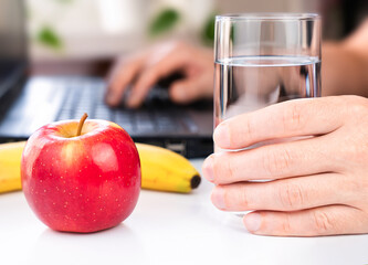 A man's hand holds a glass of water and an apple and a banana nearby. Healthy snack at work. Close-up. Selective focus.