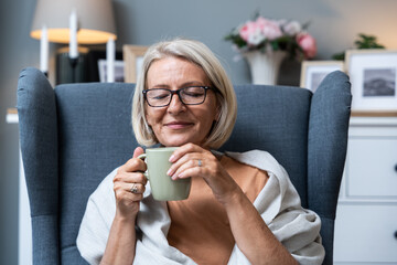Simple living. Senior woman sitting alone on chair at home drinking tea or coffee enjoying her time and life. Older female thinking about her past and happiness, satisfied with her life decisions