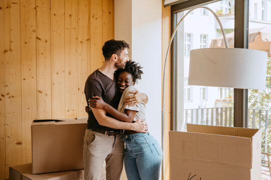 Happy couple embracing each other while standing near window at new home