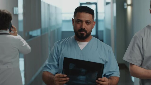 Portrait of young middle eastern doctor in scrubs examining x-ray images and looking at camera while standing in hospital corridor
