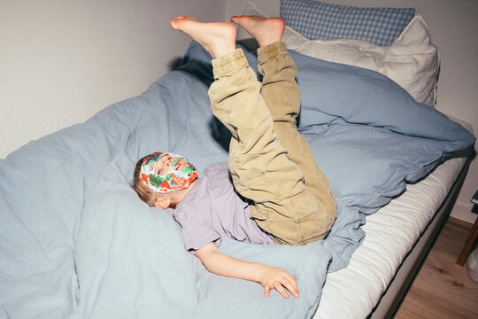 Carefree boy wearing mask while lying down on bed with legs up at home
