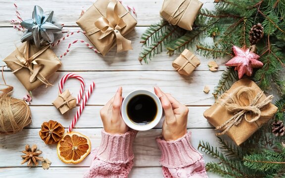 hands in pink sweater hold white cup of coffee on an old wooden work table with handmade gifts and branches of Christmas tree