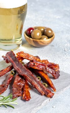 Jerky meat strips with spices, olives, rosemary and beer on wooden plate on a grey stone background