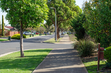The pedestrian walkway in a suburban neighborhood is lined with green trees, nature strips, and...